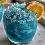 Blue Raspberry Lemonade Slushie in a chilled glass, garnished with lemon slices and mint, vibrant blue hue and frosty texture perfect for summer refreshment.