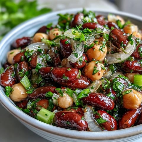 A close-up of vibrant Three-Bean Salad in a white serving bowl, featuring colorful kidney beans, chickpeas, and green beans tossed with fresh parsley and red onion.  