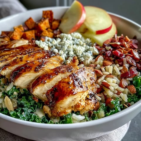 Vibrant harvest bowl with roasted chicken, sweet potatoes, kale, apples, almonds, and goat cheese, drizzled with balsamic dressing.  